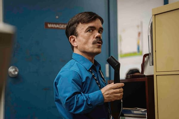 A scene from the film "ROOFMAN" featuring Peter Dinklage in a blue shirt, holding a telephone receiver, looking intently at something in an office environment.