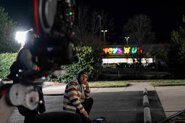 Director Derek Cianfrance on the set of the film "ROOFMAN" starring Channing Tatum and Kirsten Dunst, sitting on the ground in front of a brightly lit Toys 'R' Us storefront at night.
