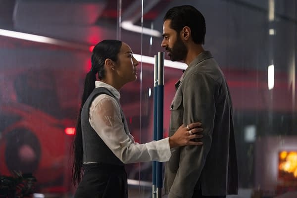 In a scene from Disney's live-action TRON: ARES, Greta Lee as Eve Kim and Hasan Minhaj as Ajay Singh are depicted in a tense moment, standing closely while facing each other against a backdrop of soft red lighting.