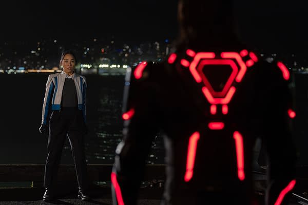 A scene from Disney's live-action TRON: ARES featuring Greta Lee as Eve Kim in futuristic clothing with blue accents, facing Jared Leto as Ares, who is in a suit with glowing red patterns, set against a nighttime city backdrop.