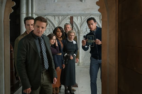 A scene from 'Wake Up Dead Man: A Knives Out Mystery' featuring seven characters, including Andrew Scott, Jeremy Renner, and Kerry Washington, standing in a stone archway with expressions of intrigue. The setting appears to be an ornate hallway with elaborate architecture.
