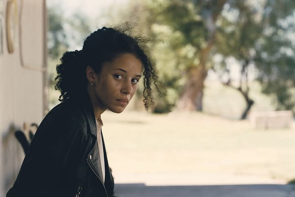 A young woman with curly hair sitting outdoors, wearing a black leather jacket and a light-colored top, gazing intently at the camera with a background of blurred greenery.