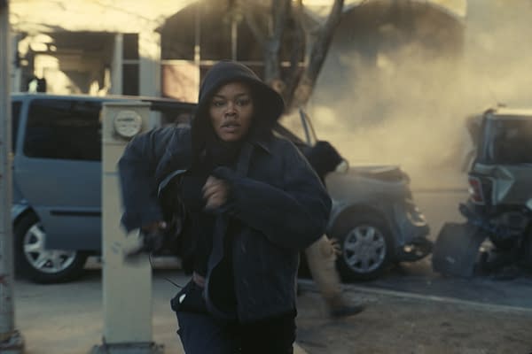 A woman in a dark hoodie runs anxiously through a chaotic scene with smoke and debris around her, suggesting a sense of urgency and danger in an urban environment.