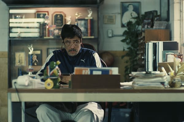 A man sits at a cluttered desk in an office filled with trophies and awards. He is wearing a dark blue athletic jacket and glasses, looking intently towards the camera.