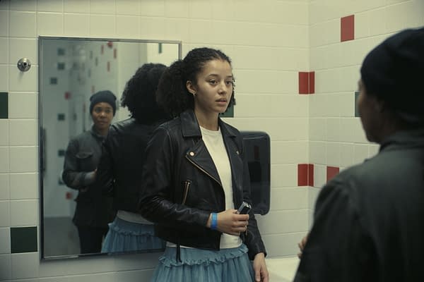 A young woman wearing a black leather jacket stands in a bathroom, looking thoughtfully at her reflection in the mirror. She holds an object in her hand, while another person is visible in the background, enhancing the scene's sense of introspection.