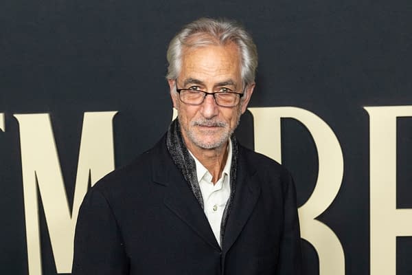 A man with gray hair and glasses is posing at a promotional event, wearing a black jacket over a white shirt, with a scarf around his neck. The background features large text, likely related to a film event.