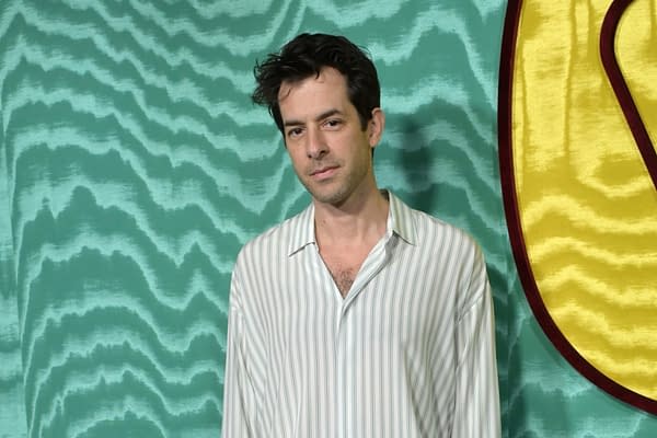 A man with tousled hair stands in front of a textured teal background. He wears a striped button-up shirt and has a neutral expression.