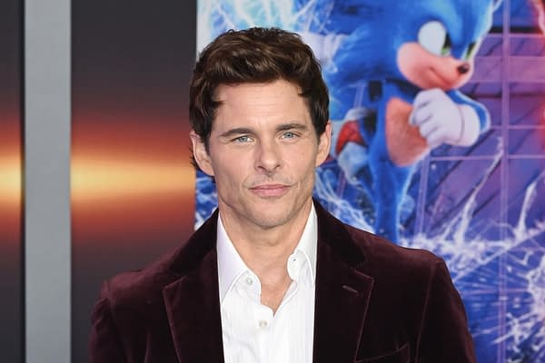 A man with short, dark hair and a well-groomed appearance wears a white shirt under a dark velvet blazer. He appears at a film premiere with a colorful background featuring a blue animated character running.