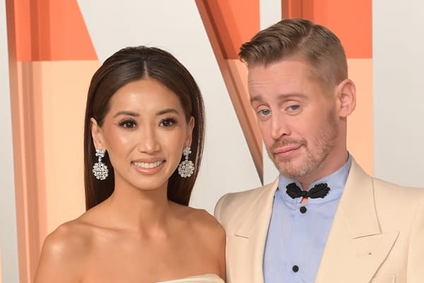 A woman with long dark hair wearing sparkling earrings and a strapless dress poses smiling next to a man in a light-colored suit with a unique collar, who has a playful expression. The background features soft geometric shapes in pastel colors.