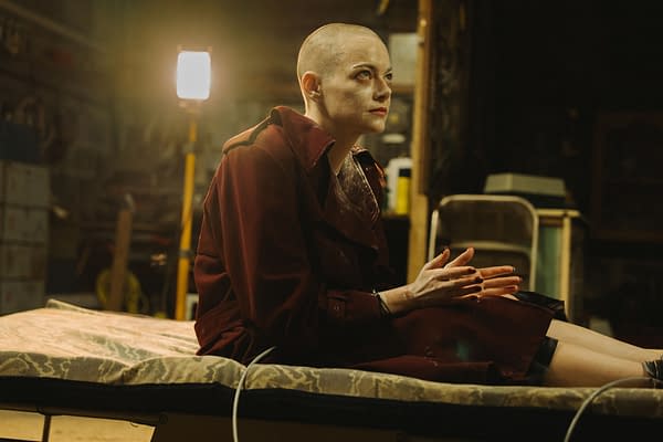 The scene from 'BUGONIA' features Emma Stone, as Michelle, with a bald head and wearing a maroon trench coat. She sits on a bed, displaying a contemplative expression in a dimly lit, industrial setting.