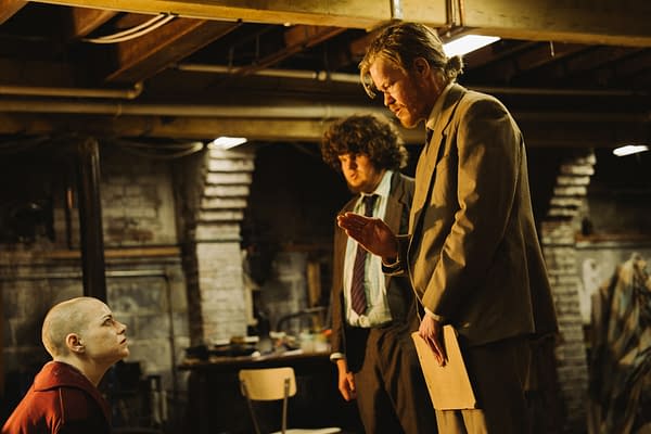 A scene from the film BUGONIA (2025) featuring three actors in a dimly lit basement setting. The individuals include a bald woman in red looking upward, a man with curly hair dressed in a suit, and another man in a suit gesturing with a folder in his hand.