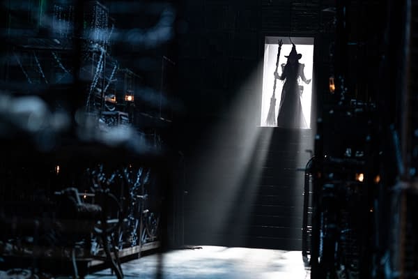 A dramatic scene from 'WICKED FOR GOOD' features a silhouette of Elphaba, portrayed by Cynthia Erivo, standing in a doorway with a broomstick. The lighting creates a mystical atmosphere, enhancing the theatrical backdrop.