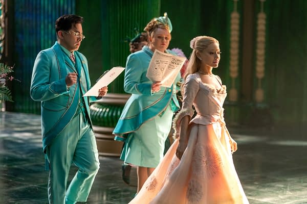 A scene from WICKED FOR GOOD featuring Bowen Yang as Pfannee, Bronwyn James as Shenshen, and Ariana Grande as Glinda, all in colorful costumes against a vibrant green background.