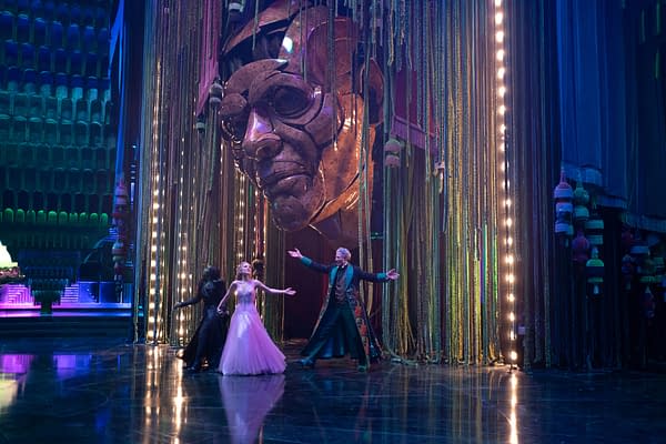 In a scene from 'Wicked: For Good,' Cynthia Erivo as Elphaba, Ariana Grande as Glinda, and Jeff Goldblum as The Wizard of Oz perform in front of a large, golden face sculpture, surrounded by colorful stage elements and dramatic lighting.