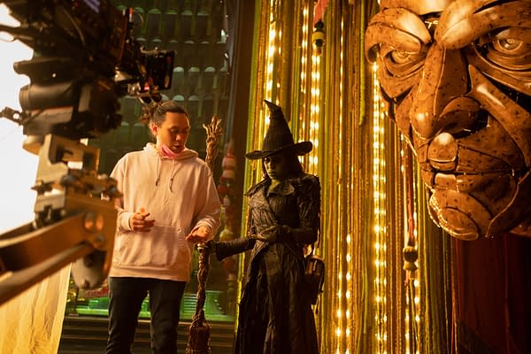 Director Jon M. Chu and actress Cynthia Erivo, dressed as Elphaba, interact on the set of 'Wicked: For Good', surrounded by bright lights and a large dramatic face prop.
