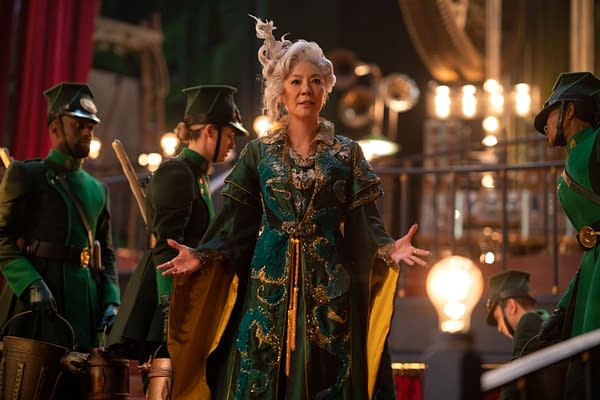 Michelle Yeoh portrays Madame Morrible in a theatrical scene from 'Wicked: For Good,' wearing an ornate green costume with gold accents, surrounded by ensemble members in green uniforms.