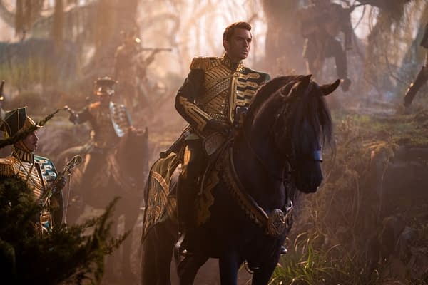 A scene from 'WICKED FOR GOOD' featuring Jonathan Bailey as Fiyero, dressed in a striking gold and green military-style costume while riding a black horse. The background includes soldiers and a misty, forested setting.