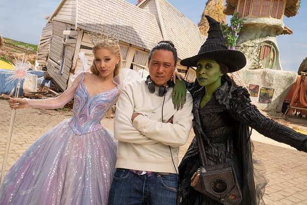 A scene from 'Wicked: For Good' showing Ariana Grande in a sparkling Glinda costume with a crown, Cynthia Erivo as Elphaba in a dark, dramatic attire, and director Jon M. Chu standing between them on a set designed to resemble a whimsical village.