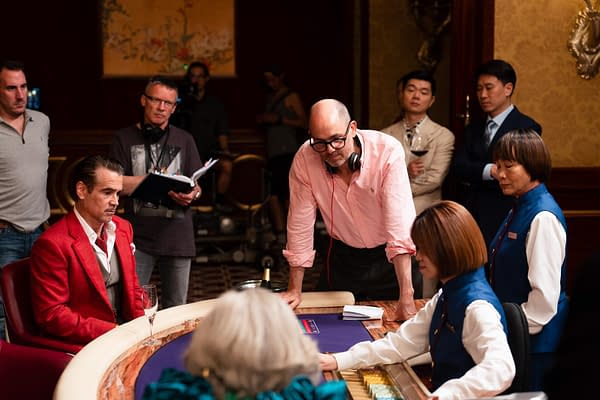 Ballad of a Small Player On the set of 'Ballad of a Small Player,' a scene is being filmed at a casino table, featuring a male lead in a red suit and crew members observing. The director, in a pink shirt and glasses, is engaged in the scene while a dealer hands over chips, and various crew members are present in the background.