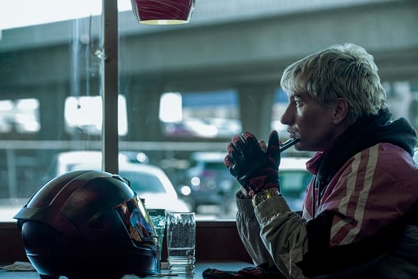 Barry Keoghan portrays a pensive character named Ormon in a scene from 'Crime 101,' sitting at a diner table with a motorcycle helmet nearby. He is dressed in a sporty jacket, wearing gloves, and gazing thoughtfully while holding a pen.