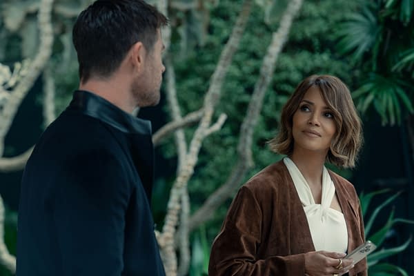 Chris Hemsworth and Halle Berry engage in a conversation amidst lush greenery, both dressed stylishly. The scene captures a moment from the project 'Crime 101,' showcasing their dynamic interaction.