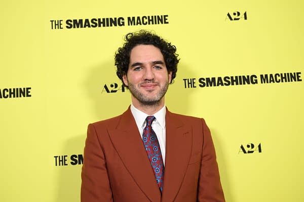 Benny Safdie poses in front of a bright yellow backdrop featuring the title "THE SMASHING MACHINE" and the A24 logo, dressed in a brown suit and patterned tie.