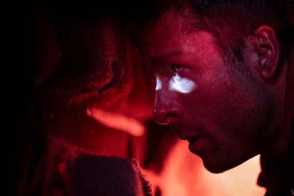 A close-up shot of a man, portrayed by Glen Powell, with intense facial expressions illuminated by a beam of light against a dark, warm-toned background, likely from the film 'The Running Man.'