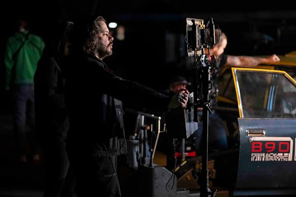 Director Edgar Wright is seen focused on a monitor while filming a scene for Paramount Pictures' 'The Running Man,' with a crew member and a vehicle partially visible in the background.
