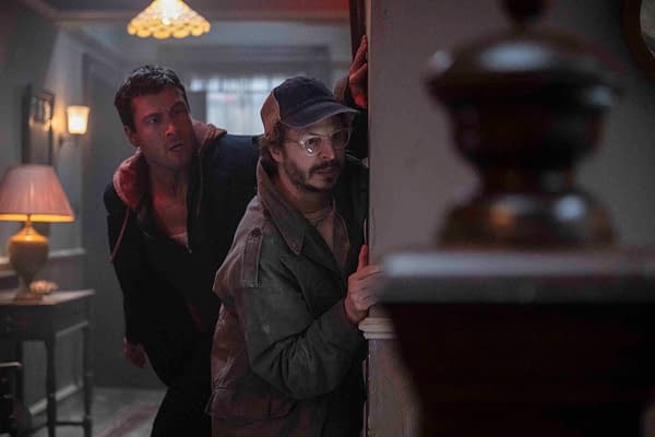 Glen Powell and Michael Cera crouch against a wall, appearing tense in a dimly lit room, conveying a sense of suspense in a scene from "The Running Man."