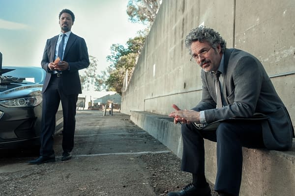 A scene from 'Crime 101' featuring two men in business attire. One stands confidently next to a car, while the other sits on a concrete ledge, appearing contemplative.