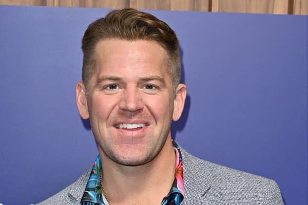 A man with short, styled hair smiles at the camera, wearing a colorful floral shirt underneath a gray blazer, with a purple background.