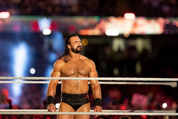A muscular male wrestler stands confidently in a wrestling ring, with his hair tied back and wearing black wrestling trunks. The background features an animated crowd, enhancing the atmosphere of a wrestling event.