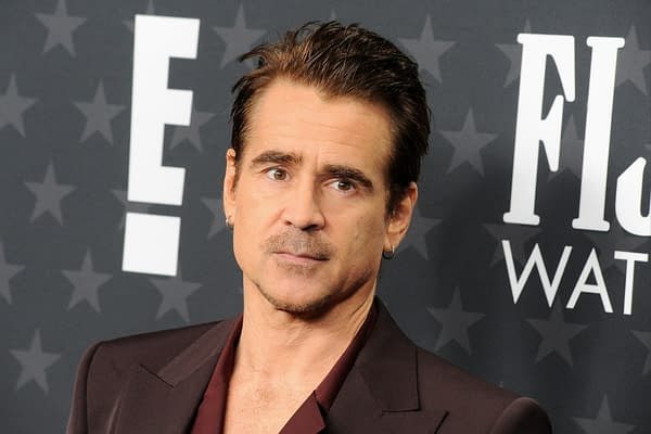 Colin Farrell is posing with a serious expression, wearing a deep burgundy suit jacket over a dark shirt, in front of a background featuring stars and the letter 'E.' This image relates to his discussion about the film Sgt. Rock.