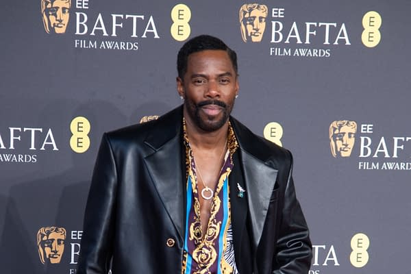 A man dressed in a stylish black leather jacket and a colorful patterned shirt poses confidently at the EE BAFTA Film Awards event, with a backdrop featuring the BAFTA logo.