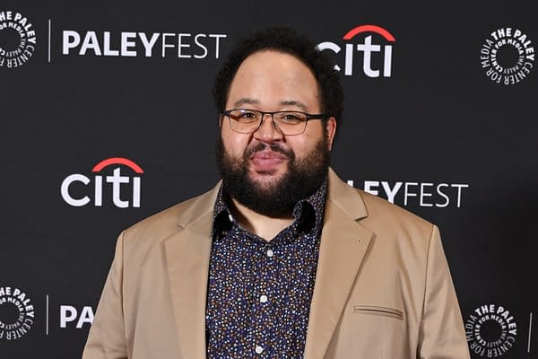 A man with curly hair and glasses smiles in front of a promotional backdrop for PaleyFest, wearing a beige blazer over a colorful floral shirt. This image is associated with the cast announcement for 'Resident Evil' featuring Zach Cherry, Kali Reis, and Johnno Wilson.