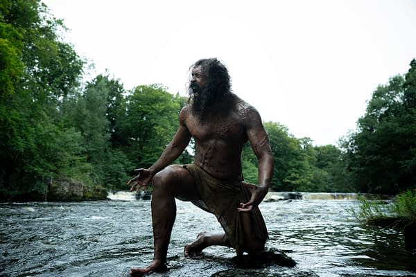 A muscular figure with long hair kneels in a river, surrounded by lush greenery, expressing intense emotion. The character appears to be weathered and adorned in a simple garment, conveying a sense of struggle or battle.