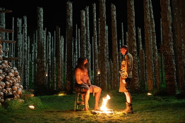 A dramatic scene set in a dark environment, featuring two men engaged in conversation around a small fire. One man sits on a stool, looking rugged and partially bare, while the other stands nearby, dressed in cargo shorts and a shirt, surrounded by tall tree-like structures and a stack of human skulls.