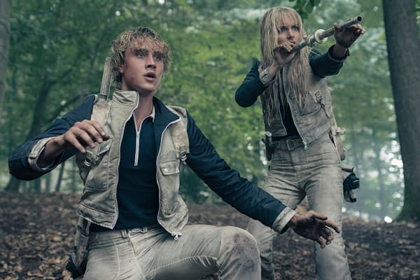 A scene from 'The Hunger Games: Sunrise on the Reaping' featuring Joseph Zada as Haymitch Abernathy, depicted in a forest setting, looking alarmed. Beside him, McKenna Grace as Maysilee Donner is poised with a weapon, both characters appear disheveled and dirtied, conveying a tense atmosphere.