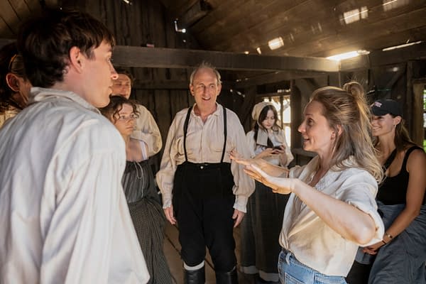 A behind-the-scenes photo from 'The Testament of Ann Lee,' featuring director Mona Fastvold engaging with the cast and crew in a rustic setting. The group is dressed in period costumes, discussing scenes while surrounded by wooden beams and warm natural light.