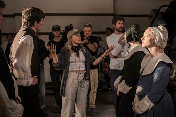A behind-the-scenes moment from 'The Testament of Ann Lee,' featuring director Mona Fastvold engaging with cast members in period costumes. The crew is gathered on set, highlighting the collaborative atmosphere of the film production.