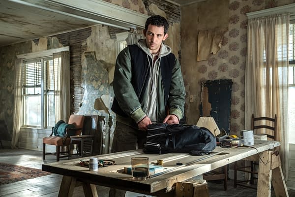 A scene from the film 'Disclosure Day,' featuring a man, Josh O'Connor, packing a black bag on a wooden table in an aged, unkempt room with floral wallpaper and a mix of furniture in the background.