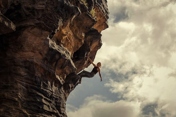 A woman, portraying Sasha in the film APEX, is seen hanging from a rocky overhang, showcasing a daring climbing pose against a backdrop of dramatic clouds. The image captures the intense and adventurous spirit of the film.