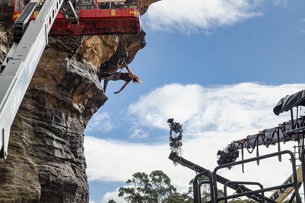 A scene from the movie 'Apex' featuring a stunt performer hanging from a rocky cliff with a film crew and equipment visible in the foreground. Bright blue sky and clouds provide the backdrop.