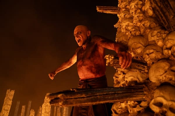 A dramatic scene featuring a bald, muscular man with a fierce expression, standing in front of a towering structure made of skulls, illuminated by an eerie glow. The man's bare torso is covered in dirt and he is gripping two wooden beams, exuding a sense of intensity and power.