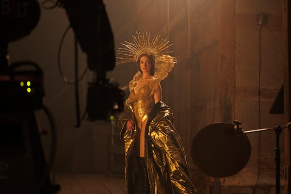 A dramatic portrait of a woman dressed in a shimmering gold outfit, featuring a large ornate headpiece with rays radiating outward. The scene is illuminated by soft lighting, creating an ethereal atmosphere.