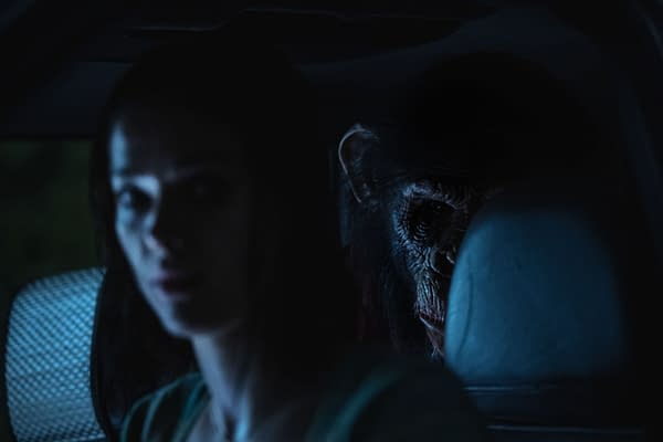 A tense scene from the film 'Primate', featuring Jessica Alexander as Hannah in a dimly lit car, with a menacing chimpanzee lurking in the background, hinting at a suspenseful atmosphere.