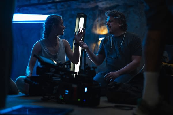 Johnny Sequoyah as Lucy engages in conversation with director Johannes Roberts on the set of the film Primate, with a focus on blue and ambient lighting.