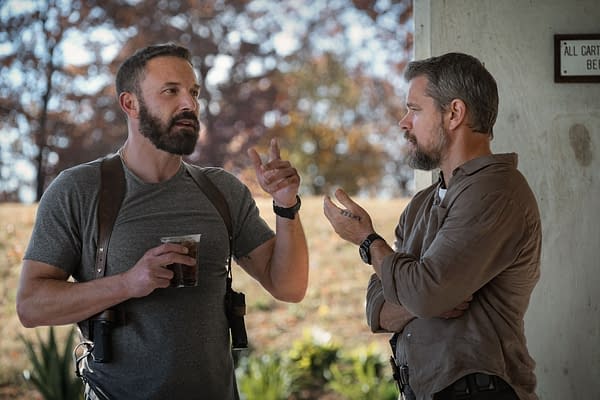 A scene from 'The Rip' featuring Ben Affleck as Detective Sergeant JD Byrne and Matt Damon as Lieutenant Dane Dumars, engaged in conversation outdoors with a blurred background of trees. Affleck is holding a drink and gesturing, while Damon listens intently.