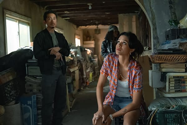 In a scene from the Netflix series 'The Rip,' Detective Mike Ro, played by Steven Yeun, stands with a firearm while Desi, portrayed by Sasha Calle, sits in a cluttered room, looking up thoughtfully. A third figure stands behind them, adding to the tense atmosphere.