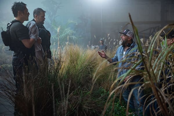 Behind-the-scenes image from 'The Rip,' featuring Steven Yeun and Matt Damon in casual attire, engaged in a discussion with writer-director Joe Carnahan on a film set surrounded by tall grass and an atmospheric backdrop.